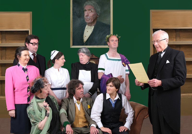 The cast of A Tomb With A View stand in front of a painting of a mad-eyed old man