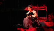 Mina sits reading a book, another on her lap. She is surrounded by red light.