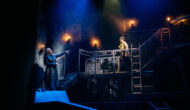 Dracula stands midway up the set pointing up at Mina who stands on the gantry above wearing a garlic flower wreath.