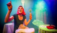The performer, Ruxy Cantor, sits at a table holding a large prop carrot. Around her are the costumes for other pickled vegetables.