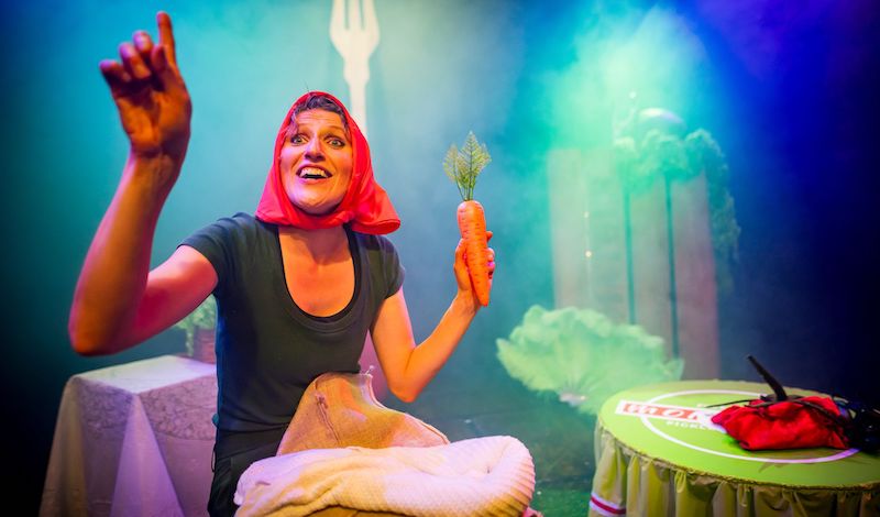 The performer sits at a table holding a large prop carrot. Around her are the costumes for other pickled vegetables.