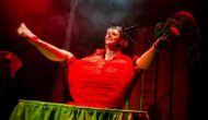 The performer, Ruxy Cantor, sits on a green table, legs akimbo, dressed as a tomato.