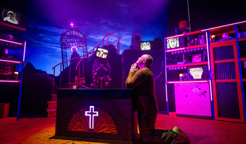 A man is on his knees praying. He is in a holy place, with marble statues, crosses and neon lights.