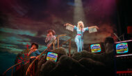 A woman dressed a country music star, with big blonde hair, a checkered jumpsuit and silver tassels is standing on a platform. Two back-up singers in cowboy hats are standing on a staircase. ‘Oppor-chancity Knocks’ is displayed on all the screens.