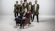 A bald older man in formal suit and casual shirt and sneakers sits in a chair. Behind him is a group of young men dressed in Parkas.