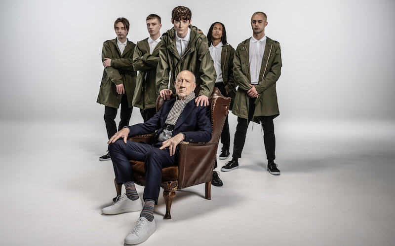 A bald older man in formal suit and casual shirt and sneakers sits in a chair. Behind him is a group of young men dressed in Parkas.