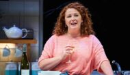 A woman with red curly hair in a pink top is in a kitchen, holding a glass of white wine.