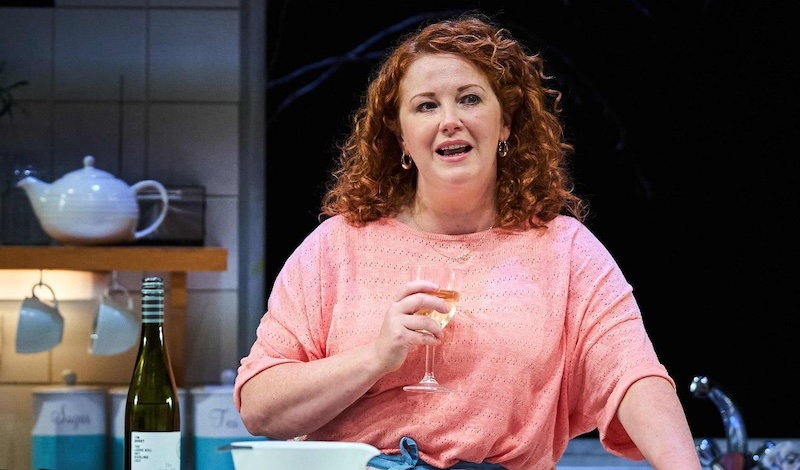 A woman with red curly hair in a pink top is in a kitchen, holding a glass of white wine.