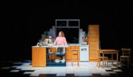 A woman with red curly hair in a pink top is in a kitchen, talking to the walls.
