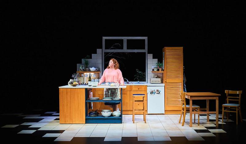 A woman with red curly hair in a pink top is in a kitchen, talking to the walls.