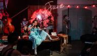 A woman with dark hair in a blue dress and silver boots sits on the edge of a small stage with a mop in her arms, pretending it is gun. Behind her, a band performs with a neon sign that says "June".