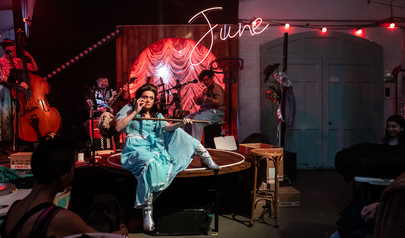 A woman with dark hair in a blue dress and silver boots sitson the edge of a smallstage with a mop in her arms, pretending it is gun. Behind her, a band performs with a neon
sign that says "June".