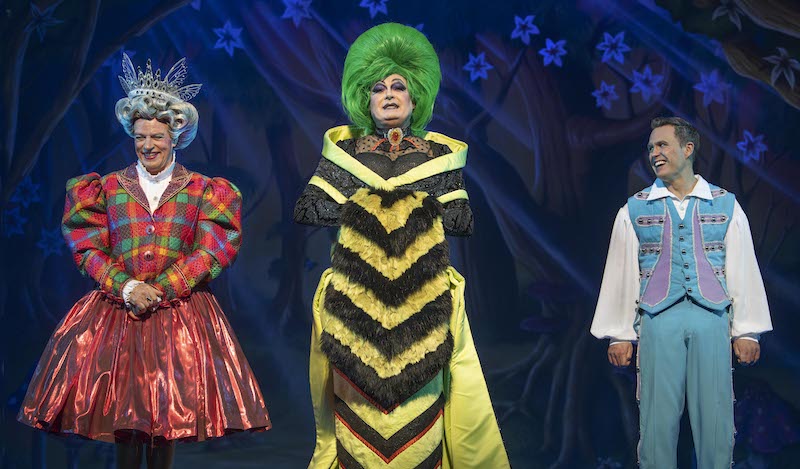 A man in a red tartan dress, a man in a wasp-like dress with a mop of green hair and a man in a blue costume stand in a line against a black background.